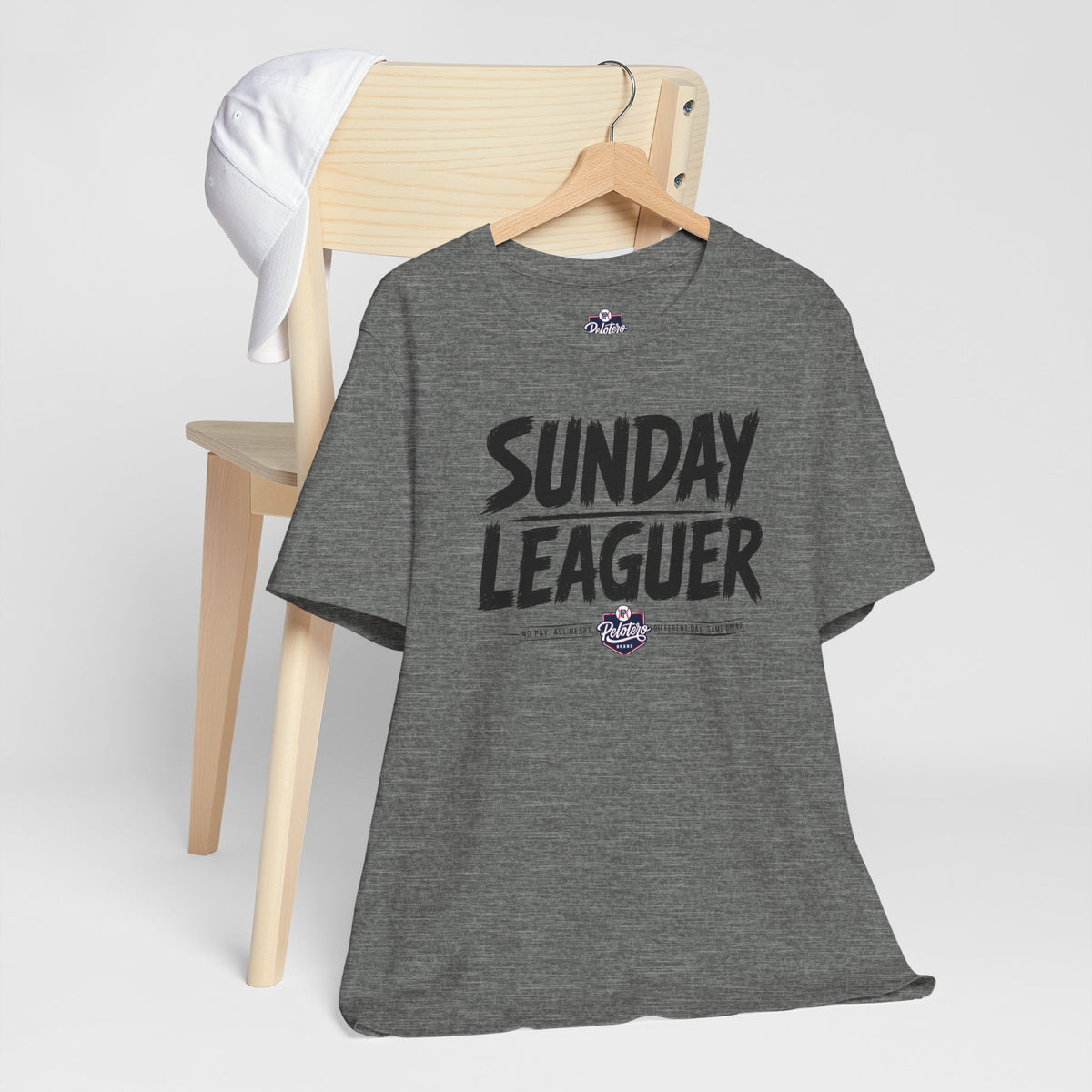 A heather gray t-shirt with 'SUNDAY LEAGUER' printed in black across the chest, hanging on a wooden hanger from a light wood chair with a white towel draped over the back, against a light gray background.
